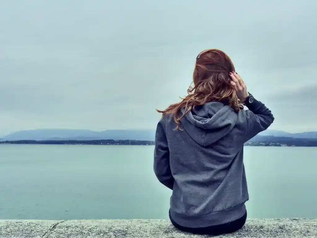 Femme vue de dos, assise face à la mer sous un ciel gris, capuche relevée, regard tourné vers l’horizon. Illustration du poème « Bats-toi » d’Hervé Gransart.