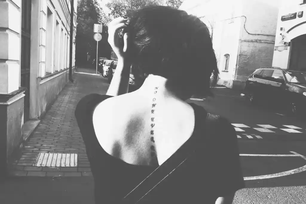 Femme vue de dos dans une rue ensoleillée, nuque dénudée et tatouage le long de la colonne vertébrale, en noir et blanc. Illustration de la nouvelle « La Belle Inconnue » d’Hervé Gransart.