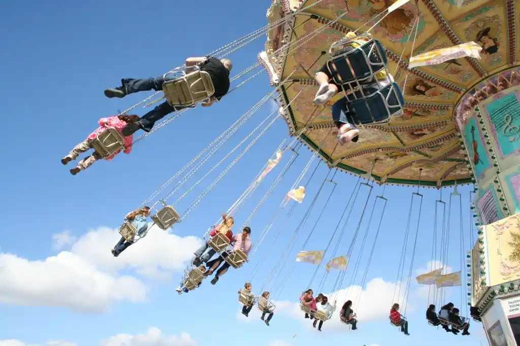 Manège de chaises volantes tournoyant sous un ciel bleu, enfants et adultes suspendus dans les airs. Représentation visuelle de la nouvelle « Le Manège » écrite par Hervé Gransart.