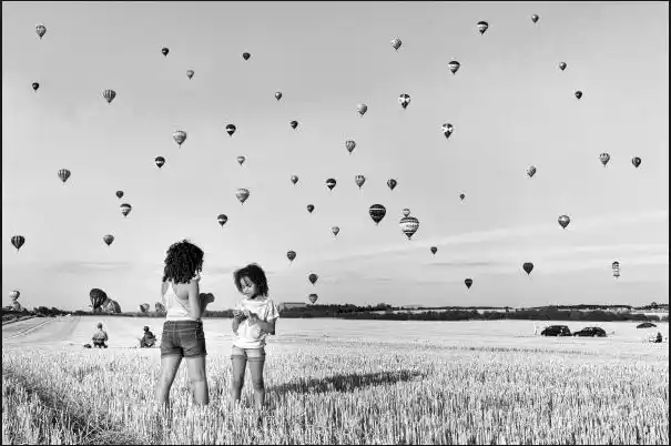 Deux jeunes filles debout dans un champ de blé, contemplant un ciel rempli de montgolfières, en noir et blanc. Illustration du poème « Sans se soucier de l’avenir » d’Hervé Gransart.