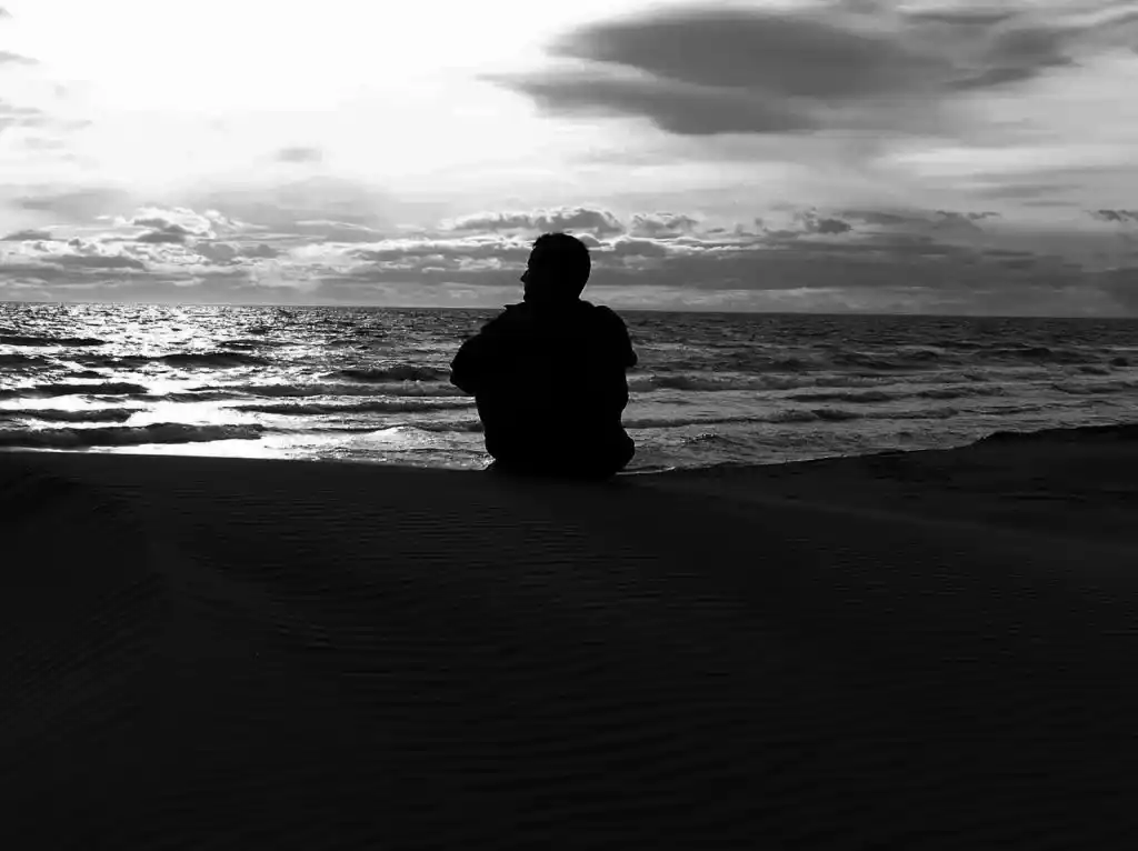 Silhouette d’un homme assis seul sur le sable face à l’océan, ciel nuageux en noir et blanc. Illustration du poème « Assis là » d’Hervé Gransart.